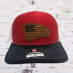 May include: A red, white and blue trucker hat with a brown leather American flag patch. The hat has a black mesh back and a white front panel. The hat has a black and white label that reads "EST. 1970 RICHARDSON. ORIGINAL 112 TRUCKER ONE SIZE".