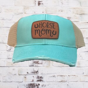 May include: A turquoise blue and brown mesh trucker hat with a brown leather patch that says "Horse Mom" in cursive.