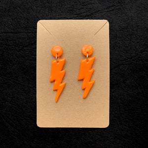 May include: A pair of vibrant orange lightning bolt earrings. Each earring has a small, round stud and a lightning bolt design. The earrings are presented on a tan card against a black backdrop.