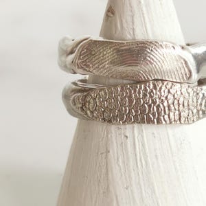 May include: Two silver rings stacked on a white cone display. The top ring features a detailed human fingerprint texture on its surface, while the bottom ring displays a textured pattern resembling small, raised scales or organic skin impressions.