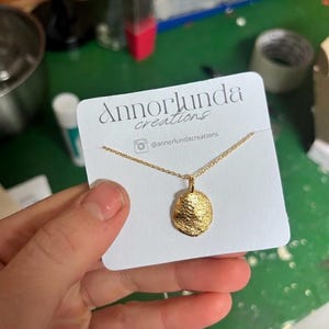 Puede incluir: Un collar de color dorado con un pequeño colgante redondo de superficie texturizada e irregular. El collar se presenta sobre una tarjeta blanca impresa con el texto Annorlunda creations y el usuario de Instagram @annorlundacreations.