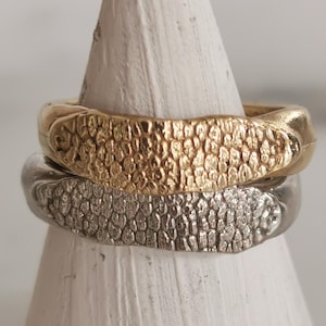 Puede incluir: Dos anillos texturizados, uno dorado y otro plateado, apilados sobre un objeto blanco en forma de cono. El anillo dorado está encima. Los anillos tienen un patrón similar a escamas.