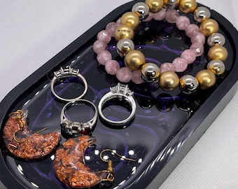 May include: A black oval tray with a purple swirl design holds a set of jewelry. The jewelry includes a pair of gold and copper crescent moon earrings, two silver rings with clear stones, and a double-strand bracelet with gold and silver beads and pink stones.