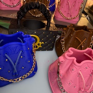 Puede incluir: Una colección de bolsos tipo cubo de colores en rosa, azul y marrón. Cada bolso presenta un cierre de cordón, una correa de cadena dorada y un patrón de logotipo en relieve. Un bolso negro tiene un asa fruncida y detalles dorados.