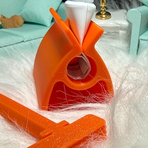 Puede incluir: Un exprimidor de tubos de plástico naranja con un tubo blanco insertado. El exprimidor tiene un diseño único con una ranura para el tubo y un asa. También se ve una herramienta naranja separada.