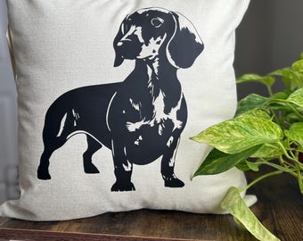 May include: A white pillow with a black silhouette of a dachshund dog printed on it. The pillow is sitting on a wooden table.