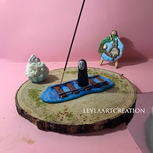 Könnte beinhalten: Ein blauer Räucherstäbchenhalter mit einer schwarzen Figur und braunen Akzenten, auf einer Holzscheibe platziert. Ein schwarzer Räucherstäbchenstab steht aufrecht. Ein kleines Gemälde und eine weiße Skulptur sind ebenfalls vorhanden. Der Text "LEYLAARTCREATION" ist sichtbar.