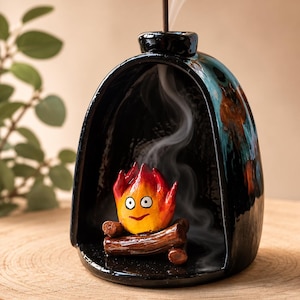 Könnte beinhalten: Ein schwarzer Räucherkegelhalter aus Keramik in Form eines gewölbten Kamins. Im Inneren sitzt eine kleine, handbemalte orange-gelbe Flammenfigur mit großen weißen Augen und einem dünnen Lächeln auf braunen Keramikscheiten, während Rauch aufsteigt.