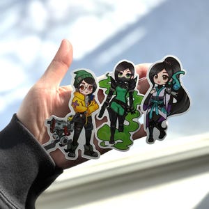 May include: Three anime-style character stickers. The first character wears a yellow jacket, glasses, and a green beanie. The second character is in a green outfit with a mask. The third character is in a white and teal outfit with long black hair.