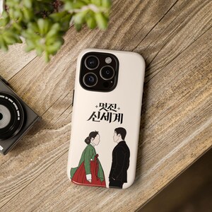 May include: A white iPhone case with a Korean drama design. The case shows two figures facing each other, with Korean text. The woman is in a green and red traditional dress, and the man wears a black suit. The case has a glossy finish.
