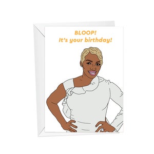 May include: A greeting card with a cartoon illustration of a woman with short blonde hair, wearing a white dress with a ruffle. The text on the card reads "BLOOP! It's your birthday!"