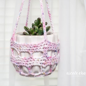 May include: A hanging plant pot with a succulent plant. The white ceramic pot is encased in a hand-crocheted holder with a pink, white, and blue speckled design. The hanger is made of the same yarn and is suspended in front of a white curtain.