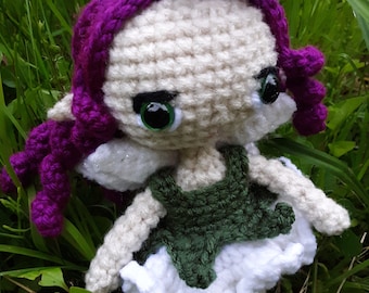 May include: A crocheted fairy doll with purple hair, a green dress, and white wings. The doll is sitting in a bed of green grass.