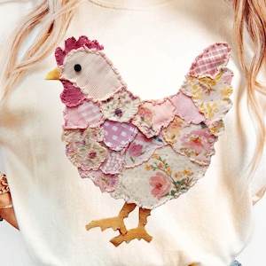 May include: Cream-colored sweatshirt featuring a patchwork chicken design in various pink floral and patterned fabrics. The chicken has a red comb, yellow beak, and brown feet. The sweatshirt is worn with blue jeans.