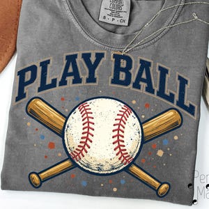 Puede incluir: Camiseta gris con las palabras "PLAY BALL" en letras mayúsculas azul marino. Debajo del texto, se muestra una pelota de béisbol y bates cruzados. La pelota es blanca con costuras rojas y los bates son de color canela.