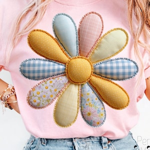 Puede incluir: Una camiseta rosa claro con un gran diseño floral colorido. La flor tiene pétalos con varios patrones, incluyendo amarillo, cuadros vichy azules e impresiones florales. El centro de la flor es amarillo con pequeños lunares. Ropa casual.