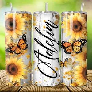 May include: Three stainless steel tumblers with lids and straws. The tumblers feature a design of sunflowers, orange butterflies, and the name "Adelin" in black script against a white wood grain background. The tumblers are on a wooden surface.