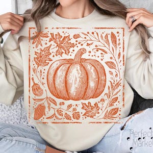 May include: Cream-colored sweatshirt featuring an orange pumpkin illustration with leaves and botanical elements. The design is framed within a rectangular border. The sweatshirt is a fall-themed apparel item.