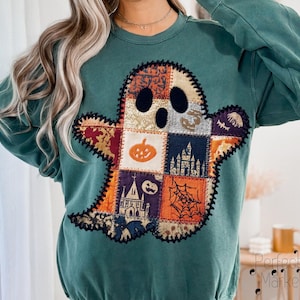 May include: Teal crewneck sweatshirt featuring a patchwork ghost design. The ghost has black eyes and a mouth, with a body made of various Halloween-themed patches, including pumpkins, spiderwebs, and haunted houses. The design is stitched onto the front.