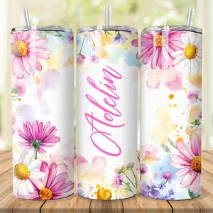 Pode incluir: Um copo branco com um design floral em aquarela com flores rosa e brancas, folhas amarelas e verdes e um fundo em aquarela rosa. O copo tem o nome "Adele" escrito em rosa na frente.