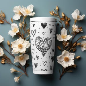 May include: White travel mug with a black and white heart pattern. The mug is surrounded by white flowers.