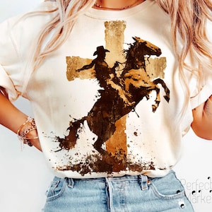 May include: Cream-colored t-shirt featuring a sepia-toned graphic of a cowboy riding a rearing horse in front of a cross. The design has a distressed, watercolor-like effect, with splatters and shading. The shirt has rolled-up sleeves.