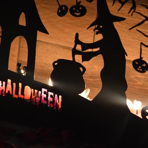 Może przedstawiać: Czarna metalowa sylwetka Halloweenowa z czarownicą mieszającą kocioł, nawiedzonym domem i napisem "HALLOWEEN" w ozdobnym kroju pisma.