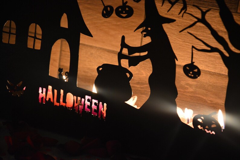 Może przedstawiać: Czarna metalowa sylwetka Halloweenowa z czarownicą mieszającą kocioł, nawiedzonym domem i napisem "HALLOWEEN" w ozdobnym kroju pisma.