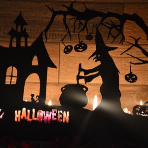 Może przedstawiać: Czarna metalowa dekoracja halloweenowa z czarownicą mieszającą kocioł, nawiedzonym domem i dynią z wyciętym uśmiechem. Dekoracja ma wycięcia na świece, a na dole napisano "HALLOWEEN".