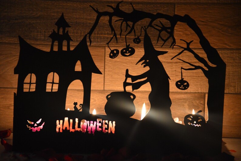 Może przedstawiać: Czarna metalowa dekoracja halloweenowa z czarownicą mieszającą kocioł, nawiedzonym domem i dynią z wyciętym uśmiechem. Dekoracja ma wycięcia na świece, a na dole napisano "HALLOWEEN".