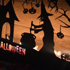 Może przedstawiać: Czarna metalowa dekoracja halloweenowa z czarownicą mieszającą kocioł, nawiedzonym domem i napisem "HALLOWEEN" w przerażającym stylu. Dekoracja jest oświetlona świecami.