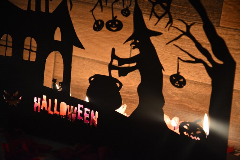 Może przedstawiać: Czarna metalowa dekoracja halloweenowa z czarownicą mieszającą kocioł, nawiedzonym domem i napisem "HALLOWEEN" w przerażającym stylu. Dekoracja jest oświetlona świecami.