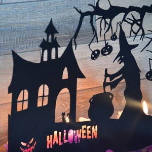 Może przedstawiać: Czarna metalowa dekoracja halloweenowa przedstawiająca sylwetkę domu, czarownicę mieszającą kocioł i drzewo z wiszącymi dyniami. Słowo "HALLOWEEN" jest napisane kolorowymi literami na dole.