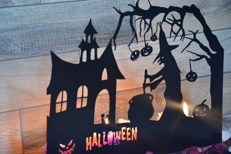 Może przedstawiać: Czarna metalowa dekoracja halloweenowa przedstawiająca sylwetkę domu, czarownicę mieszającą kocioł i drzewo z wiszącymi dyniami. Słowo "HALLOWEEN" jest napisane kolorowymi literami na dole.