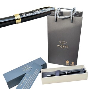 May include: A black Parker IM ballpoint pen with a silver trim and a gold inscription that reads "Dream, Write, Achieve". The pen is in a white box with a blue lid and a Parker logo. The pen is also in a gray shopping bag with a Parker logo.