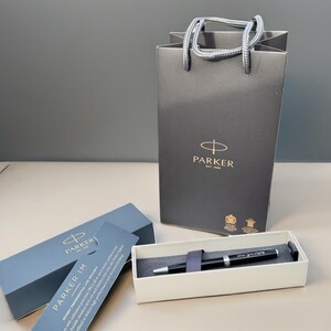 May include: A black Parker IM pen in its box with a Parker shopping bag in the background. The pen has a silver trim and the Parker logo is visible on the pen and the bag.