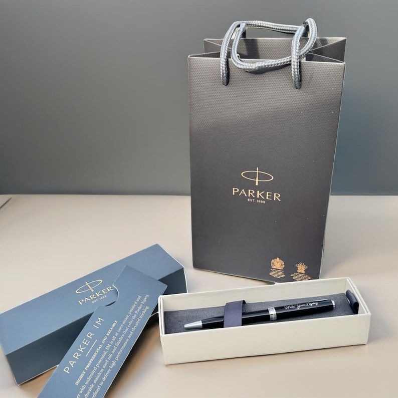 May include: A black Parker IM pen in its box with a Parker shopping bag in the background. The pen has a silver trim and the Parker logo is visible on the pen and the bag.
