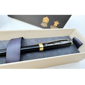 May include: A black pen with gold accents in a cream-colored box. The pen has the words "Dream, Write, Achieve" engraved on it.