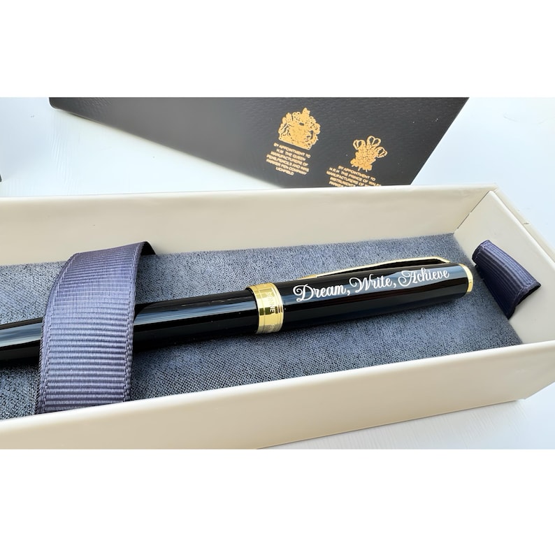 May include: A black pen with gold accents in a cream-colored box. The pen has the words "Dream, Write, Achieve" engraved on it.