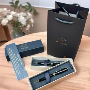 May include: A black Parker IM ballpoint pen in a white box with a black ribbon. The pen has gold accents and the Parker logo is visible on the pen and the box. The box is sitting on a wooden surface. The pen is in a gift bag with the Parker logo.