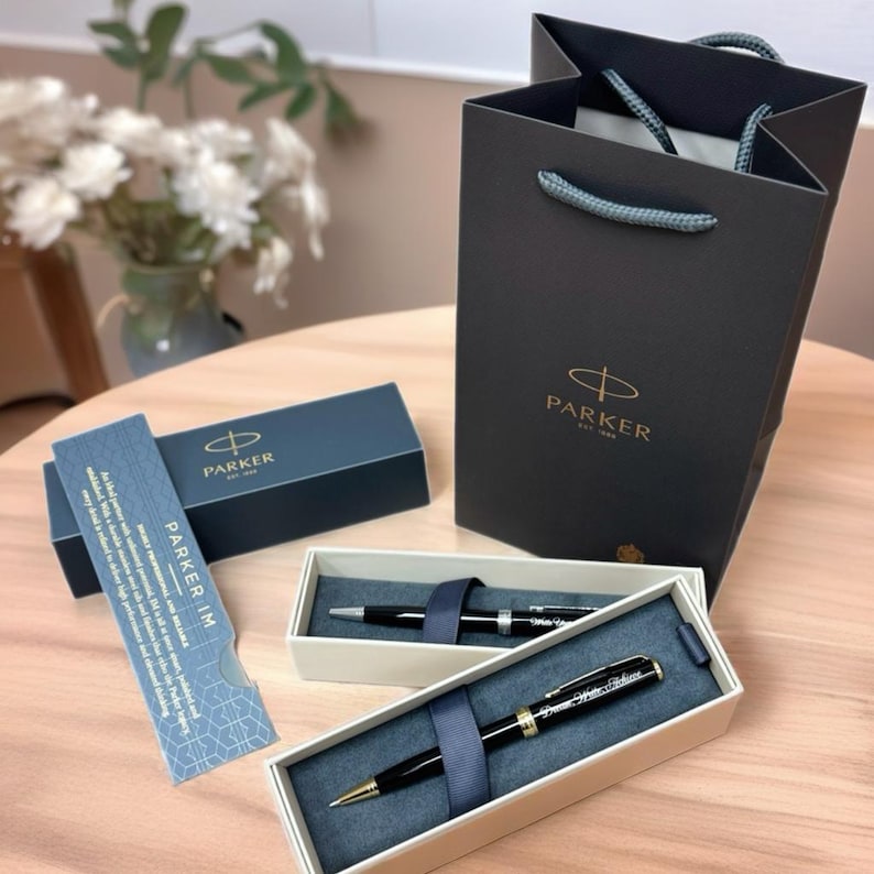 May include: A black Parker IM ballpoint pen in a white box with a black ribbon. The pen has gold accents and the Parker logo is visible on the pen and the box. The box is sitting on a wooden surface. The pen is in a gift bag with the Parker logo.