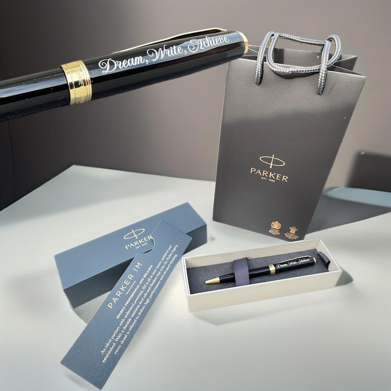 May include: A black Parker IM ballpoint pen with gold accents and the inscription "Dream, Write, Achieve" in a black gift box with a white tag. The pen is also shown in a black Parker gift bag.