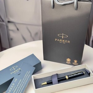 May include: A black Parker IM ballpoint pen with gold accents and a personalized inscription that reads "Dream, Write, Achieve". The pen is in its box, which is lined with gray fabric. The pen is a writing instrument.