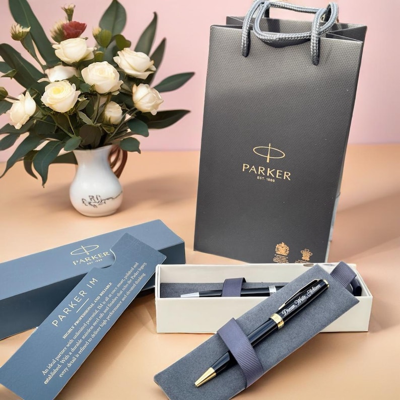 May include: A black Parker IM ballpoint pen with gold accents and a personalized inscription in a white box with a gray velvet lining. The pen is next to a black gift bag with the Parker logo.