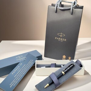 May include: A black Parker IM ballpoint pen with a gold trim and a personalized inscription. The pen is in a gray velvet pen case and is packaged in a white box with a black Parker logo. The pen is also included in a black Parker shopping bag.