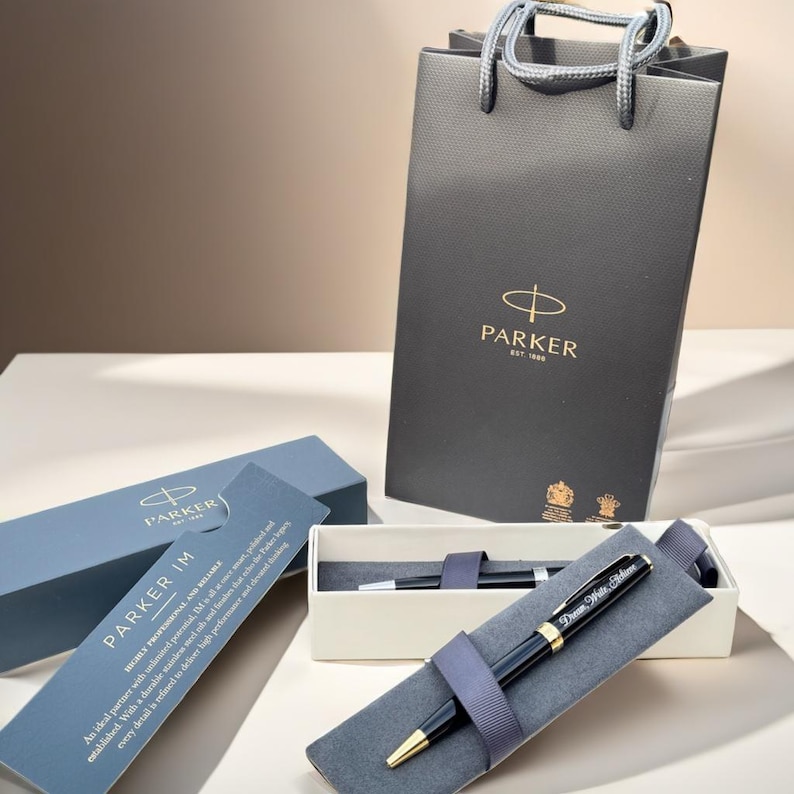 May include: A black Parker IM ballpoint pen with a gold trim and a personalized inscription. The pen is in a gray velvet pen case and is packaged in a white box with a black Parker logo. The pen is also included in a black Parker shopping bag.