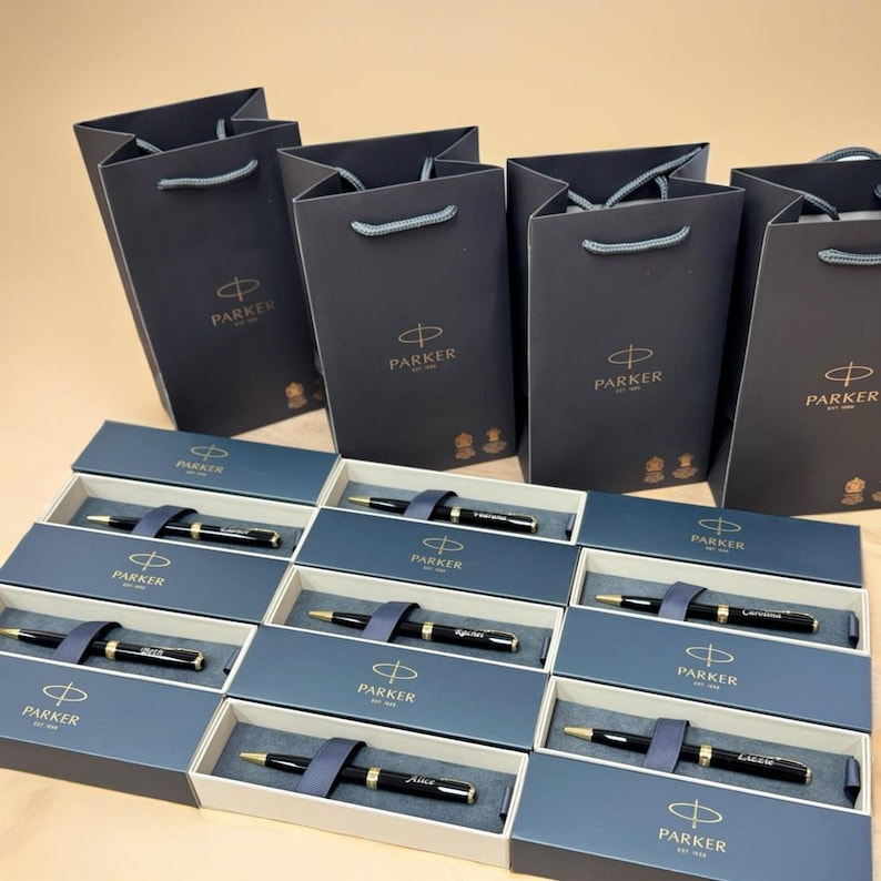 May include: A collection of black Parker pens in individual boxes with personalized names. The boxes are arranged in rows on a light brown surface.  The pens are packaged in black gift bags with gold Parker logos.