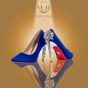 May include: A pair of blue suede high heels with a red sole and a jeweled embellishment on one shoe. The shoes are reflected in a shiny surface. The brand name "OORVI DESAI London since 2003" is visible in the background.