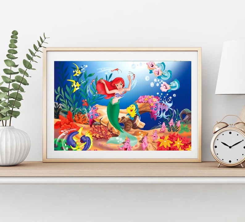 Puede incluir: Una ilustraci&oacute;n enmarcada de Ariel, la Sirenita, nadando en el oc&eacute;ano con peces y corales coloridos. Lleva su caracter&iacute;stico sujetador de concha p&uacute;rpura y su cola verde.