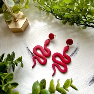 May include: A pair of red snake-shaped dangle earrings with a round red accent at the top of each earring.